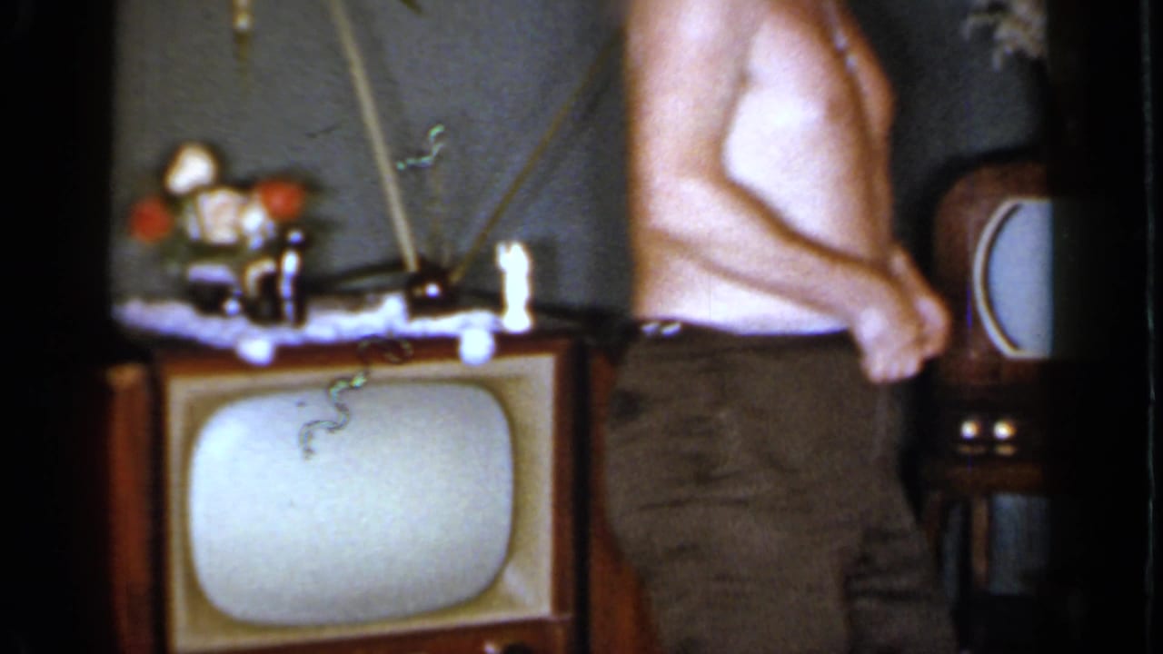 The man stands partly turned in a Mineola living room, with a wood television and starburst wall clock visible behind him.