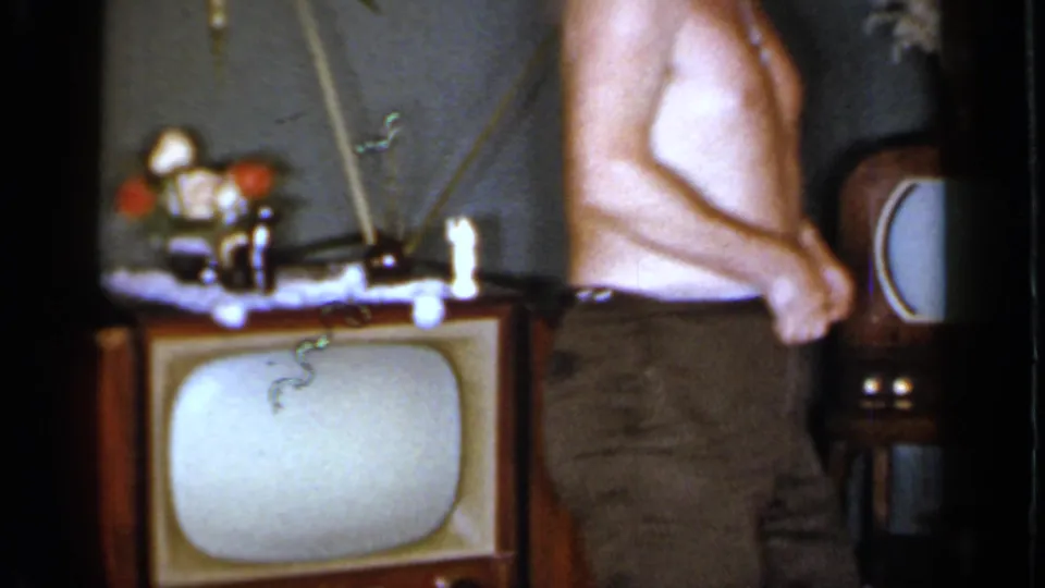 The man stands partly turned in a Mineola living room, with a wood television and starburst wall clock visible behind him.