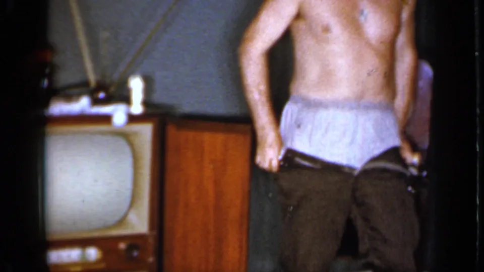 A shirtless adult man pulls down his trousers in a 1966 Mineola living room, with a vintage television at the side.