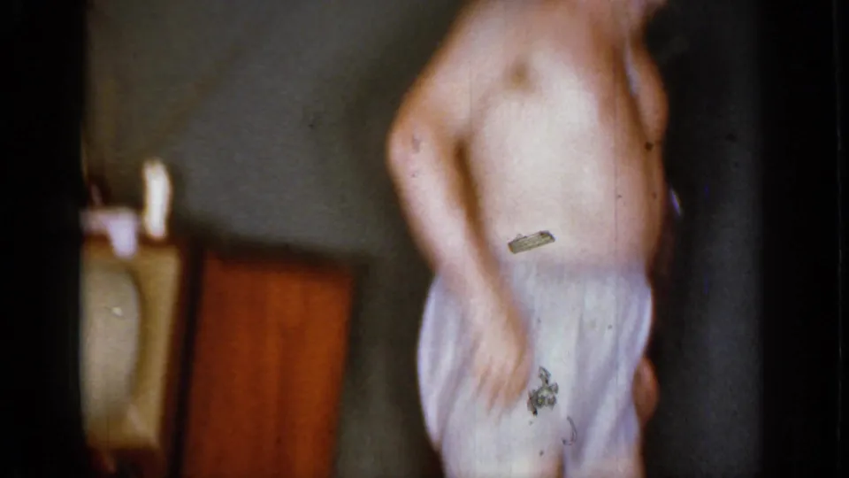 In a 1966 Mineola home-movie frame, a shirtless man in light boxers dances in a dim private interior.