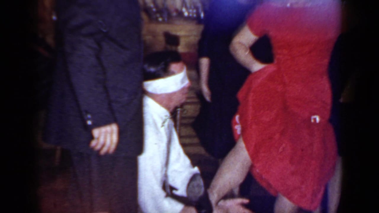 Blindfolded man kneels beside a woman in a red dress during an indoor party.