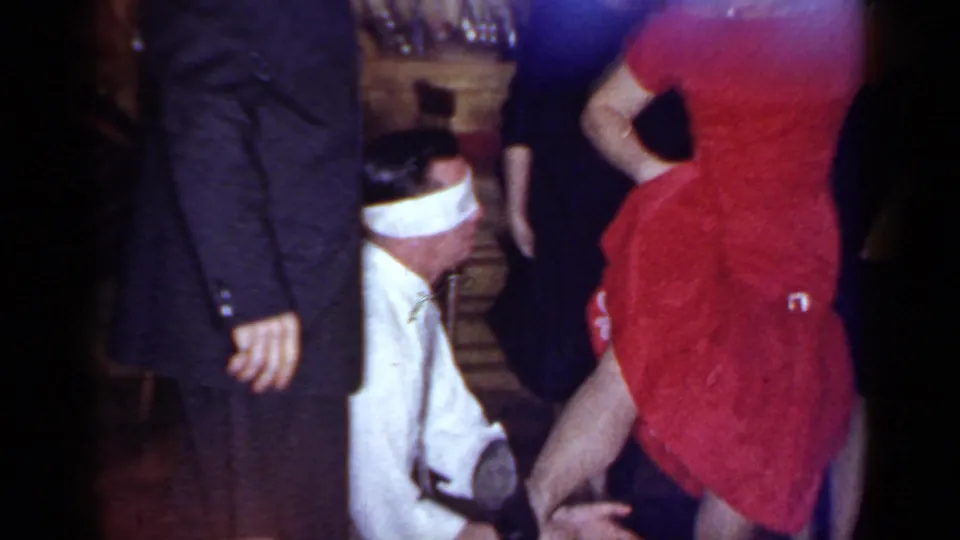 1966 Mineola home-movie frame showing a blindfolded man kneeling near a woman in a red dress as guests stand nearby.