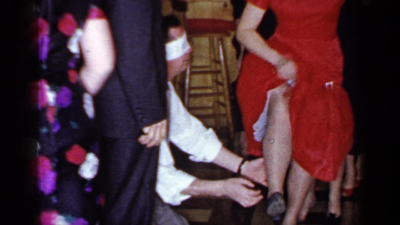 Blindfolded man reaches toward a woman in a red dress while party guests stand around him.