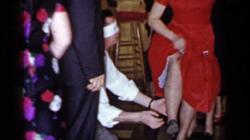 1966 Mineola party frame with a blindfolded man kneeling and reaching toward a woman in a red dress among standing guests.
