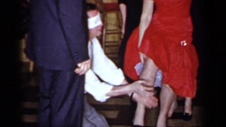 1966 Mineola home-movie frame showing a blindfolded man crouched by the legs of a woman in a red dress during a party game.