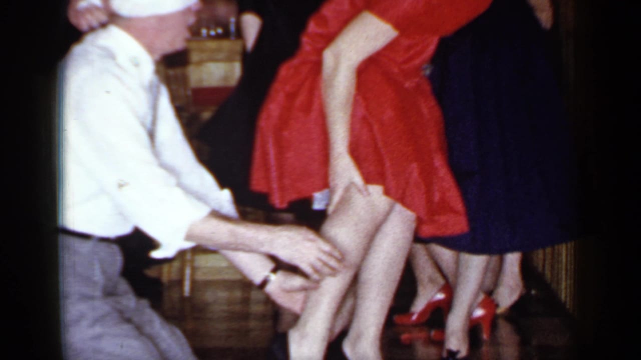 Blindfolded man kneels near women in dresses during an indoor party game.