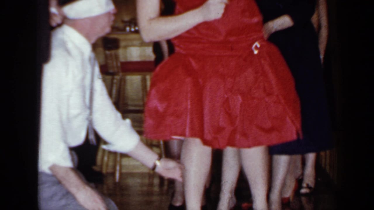 Blindfolded man reaches toward a woman in a red dress as others stand nearby.
