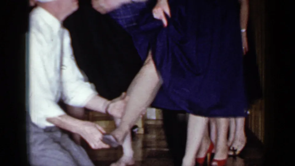 A blindfolded man kneels near a woman in a purple dress, with other party guests partly visible in the Mineola interior.