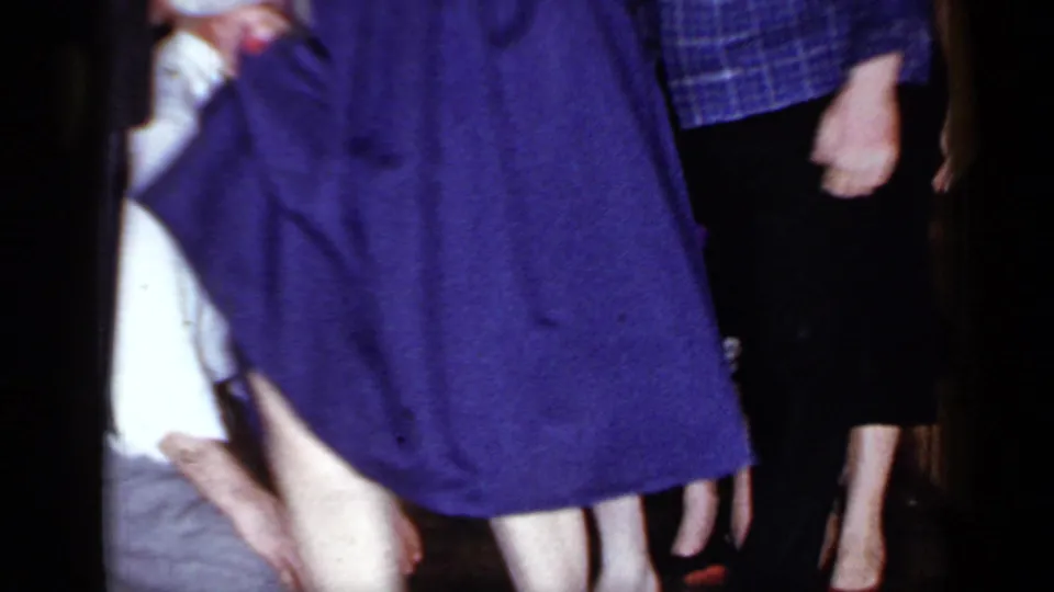 The frame holds close on a woman in a purple dress with the kneeling blindfolded man partly visible at the edge of the party scene.