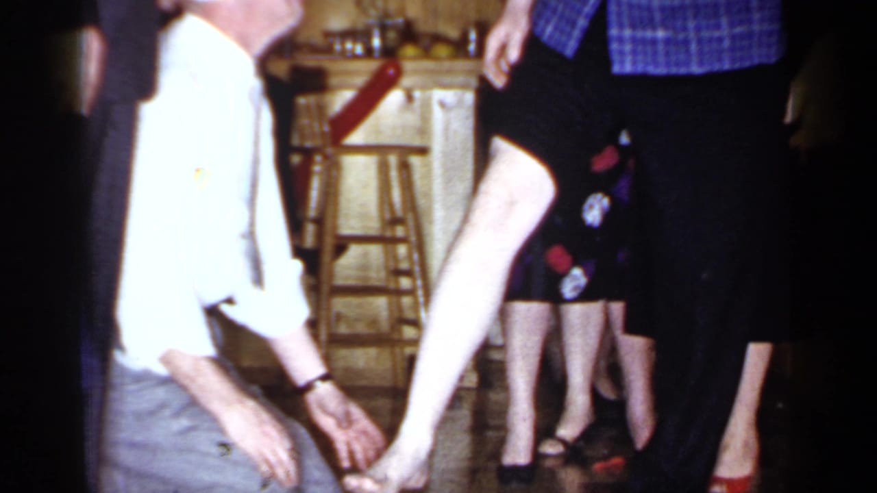 Kneeling man reaches toward a woman's leg beside adults standing in a kitchen.