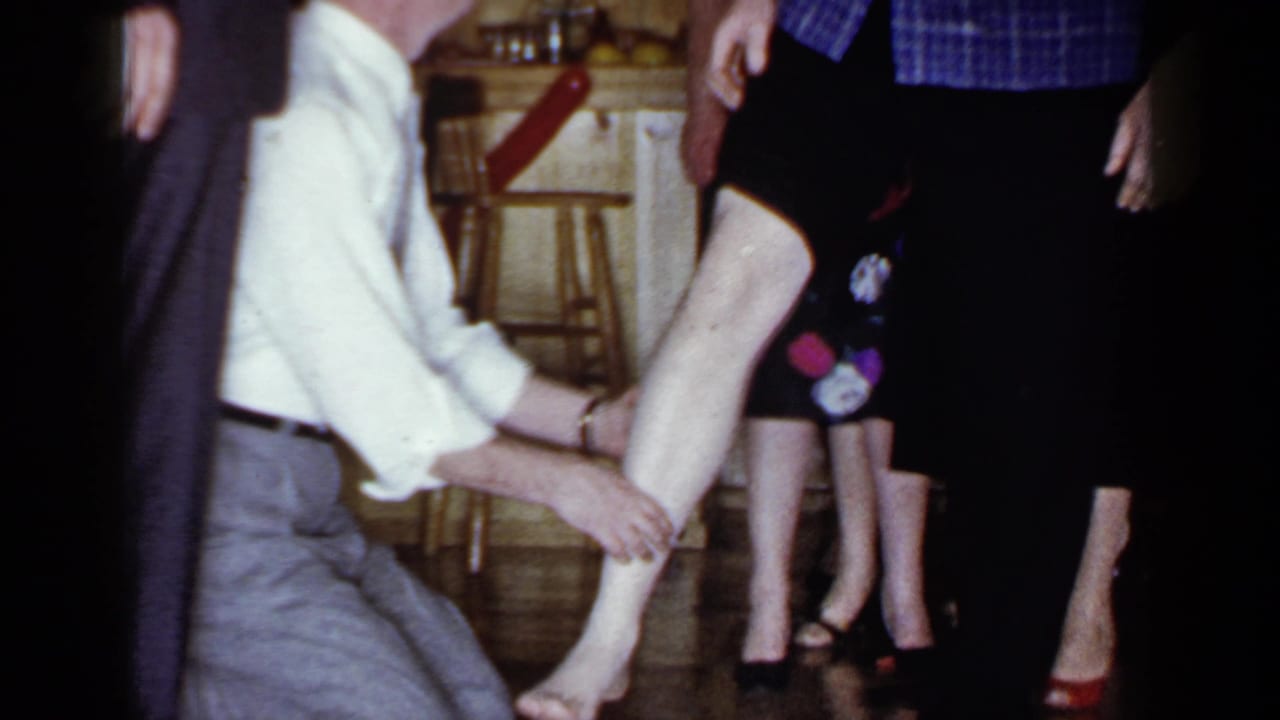 The kneeling man holds a woman's lower leg in a Mineola kitchen setting, with other adults partly visible around him.
