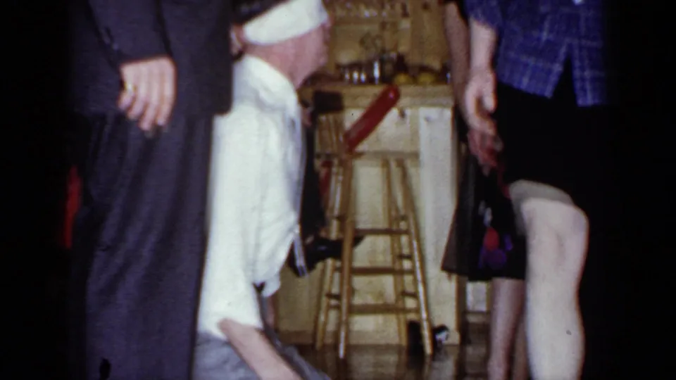 A partially blocked 1966 home-movie frame shows adults in a kitchen while the kneeling man remains near the woman's legs.