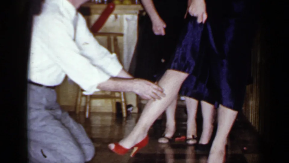 In the Mineola kitchen, the kneeling man touches a woman's leg as she stands in red heels beside another adult in dark clothing.