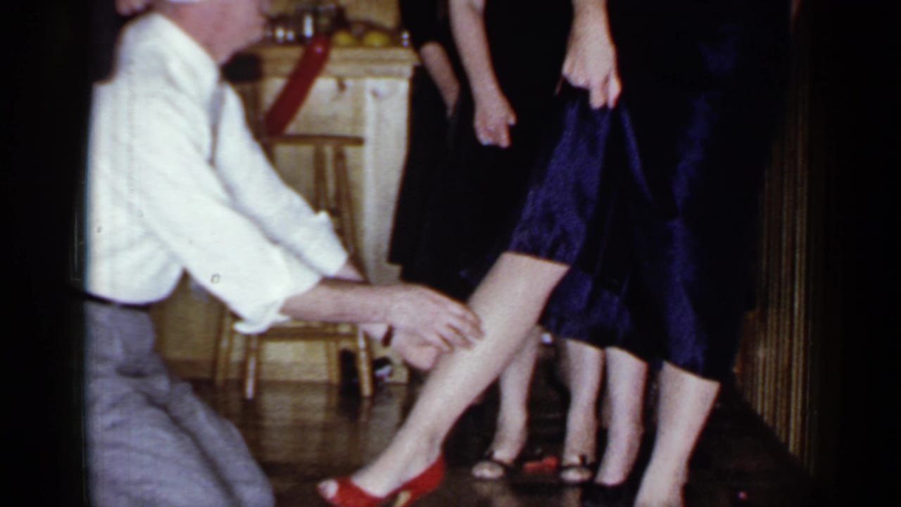 Blindfolded man kneels beside a woman in a dark dress and red heels during a party game.