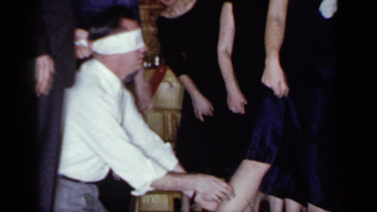 Blindfolded man reaches toward a woman's ankle while adults in dark clothing stand around him.