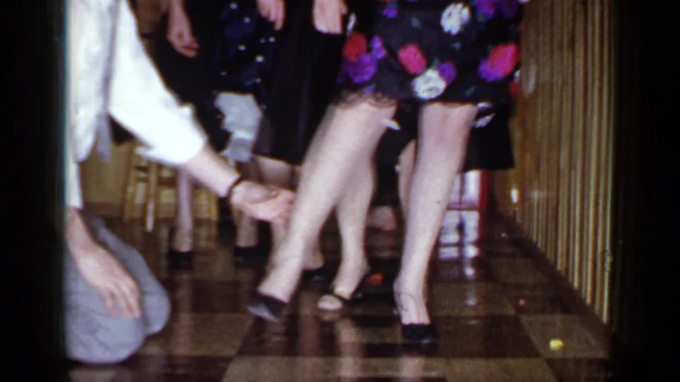 The home-movie frame shows the blindfolded party game from a low angle, with women's heels and a floral dress prominent in the room.