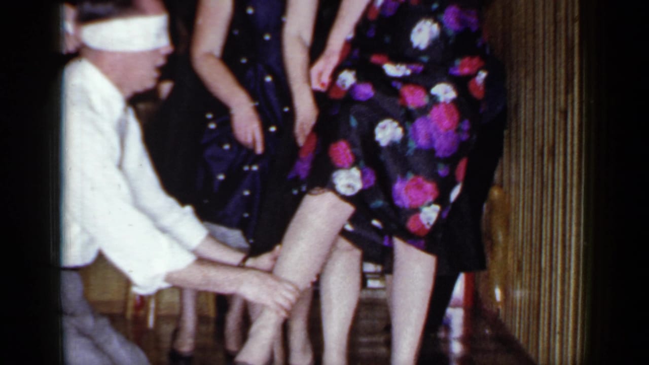 Blindfolded man kneels beside women standing in dresses on an indoor staircase or landing.