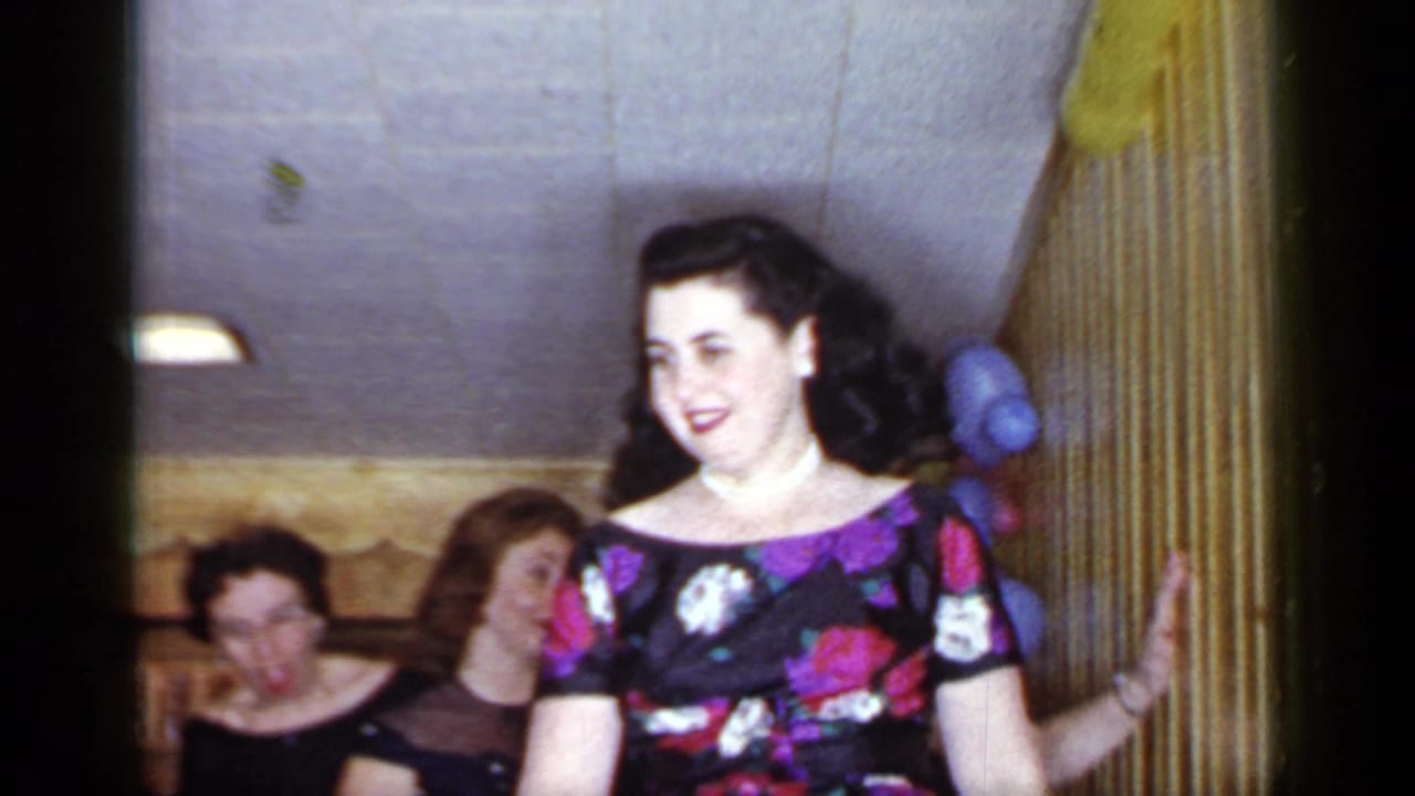 A woman in a floral dress smiles in a private Mineola interior during the 1966 party-game footage.