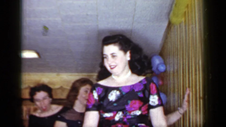 A woman in a floral dress smiles in a private Mineola interior during the 1966 party-game footage.