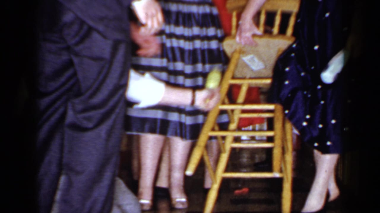 Blindfolded man reaches toward a wooden chair while adults stand nearby.