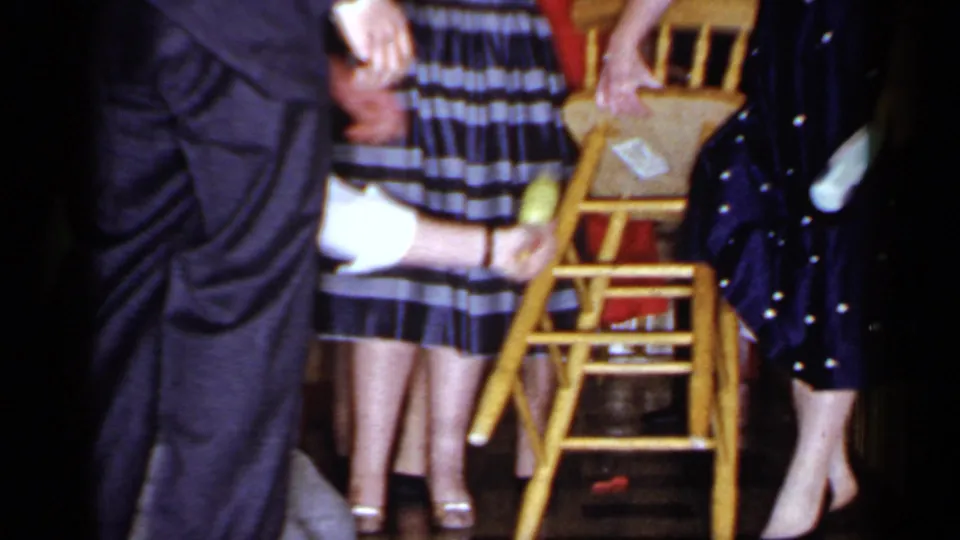 The blindfolded man reaches toward a wooden chair as surrounding adults guide or watch the Mineola party game.