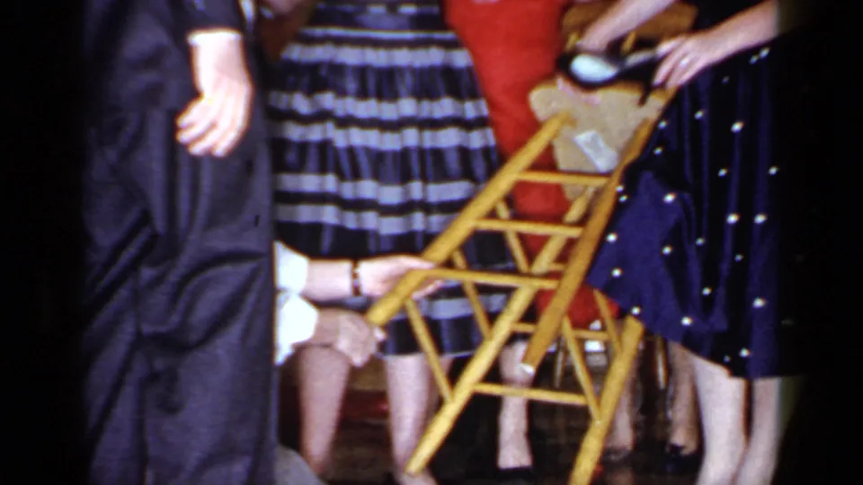 Guests in patterned dresses hold and adjust a wooden chair while the blindfolded participant reaches upward during the party game.