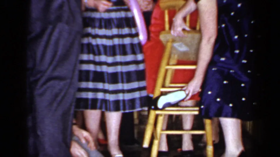 A woman in a dark polka-dot dress holds the wooden chair as the blindfolded man reaches below it in the 1966 party-game scene.