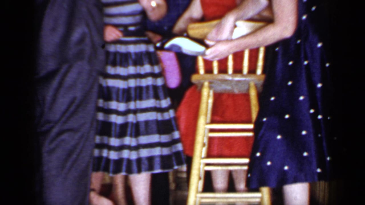 Adults at an indoor party gather around a wooden chair, with patterned dresses and dark suits visible.