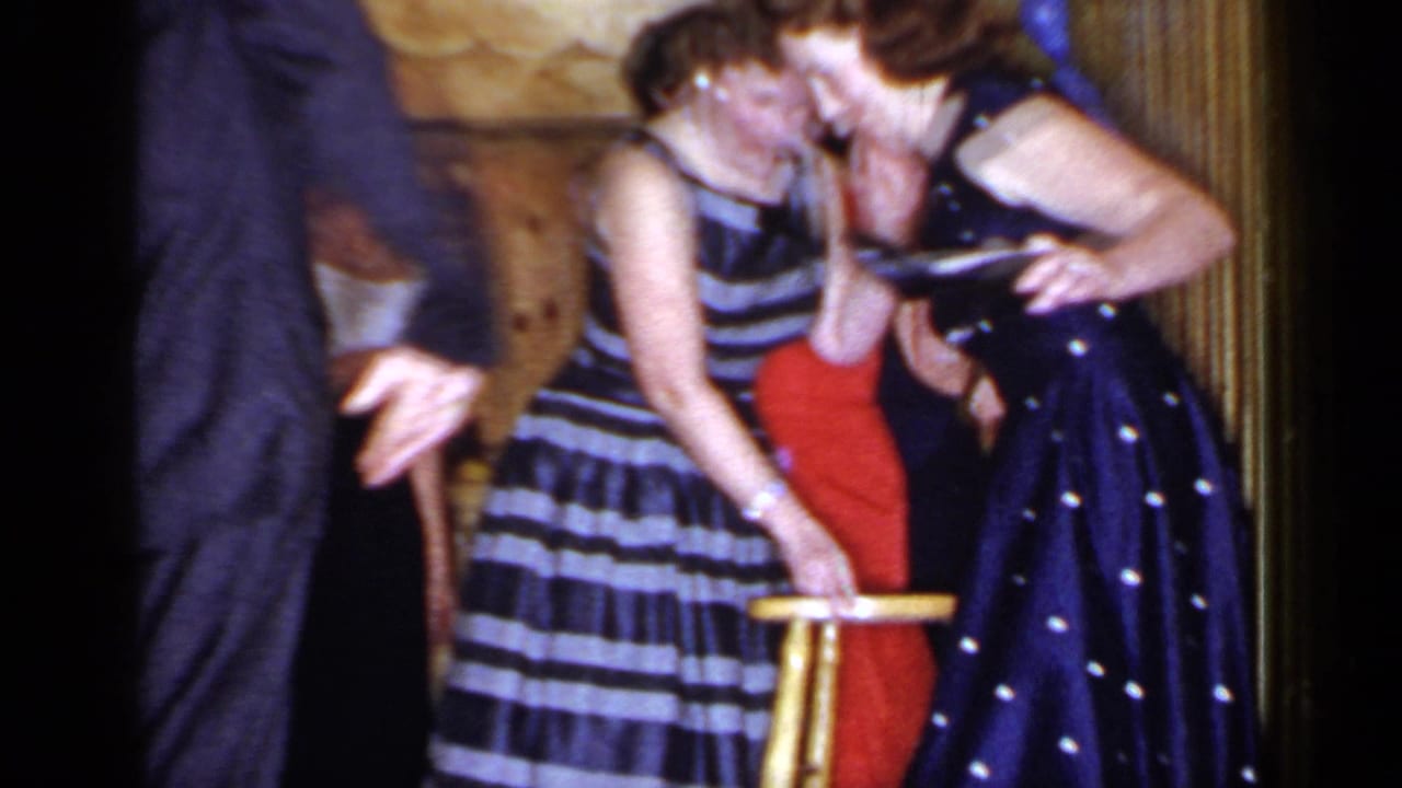 Women lean toward a wooden chair as other party guests stand close by in a warm interior.