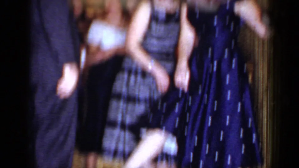 Motion-blurred 8mm frame of adults moving around a chair during a 1966 Mineola party game in a private room.