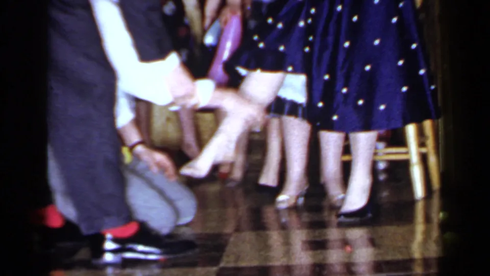 1966 Mineola party frame with a seated woman, nearby guests, dress shoes, patterned dresses, and a wooden chair in a private interior.