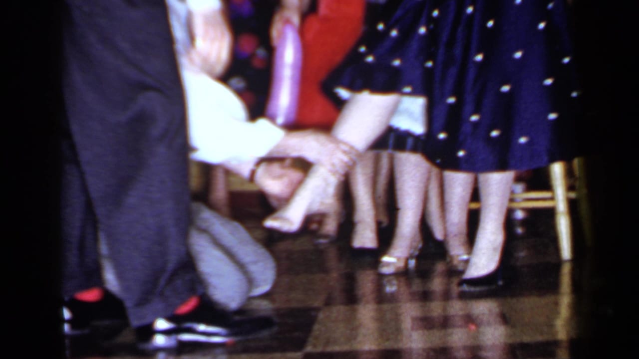 Man kneels near a woman's raised foot as party guests stand around them.