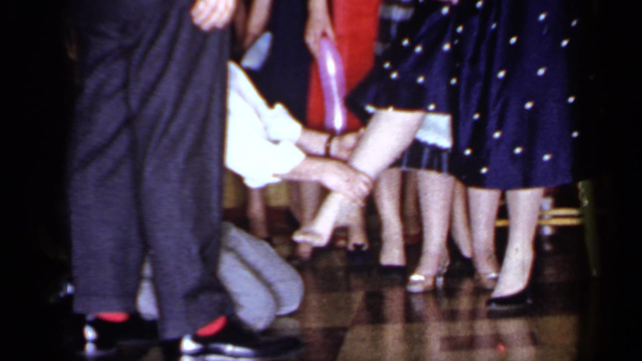Kneeling man holds a woman's foot while guests stand close by on a shiny floor.