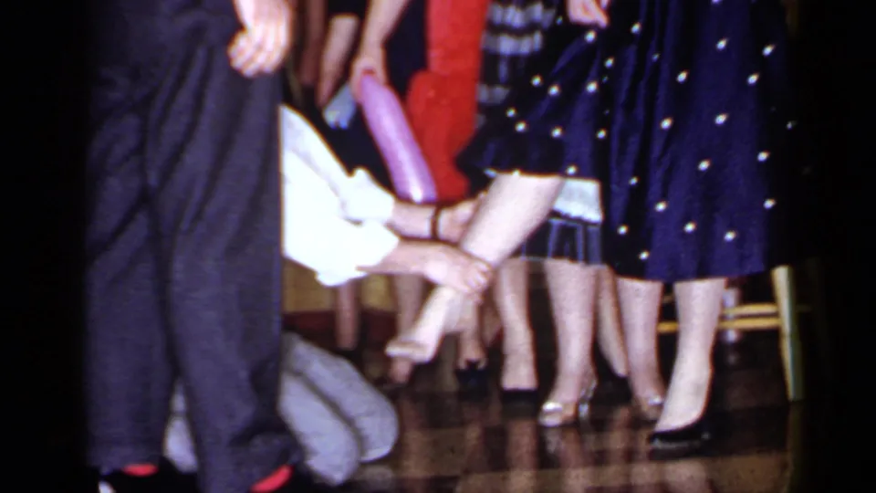 The 8mm frame shows the kneeling man at floor level near the woman's foot, with period dresses and shoes filling the background.