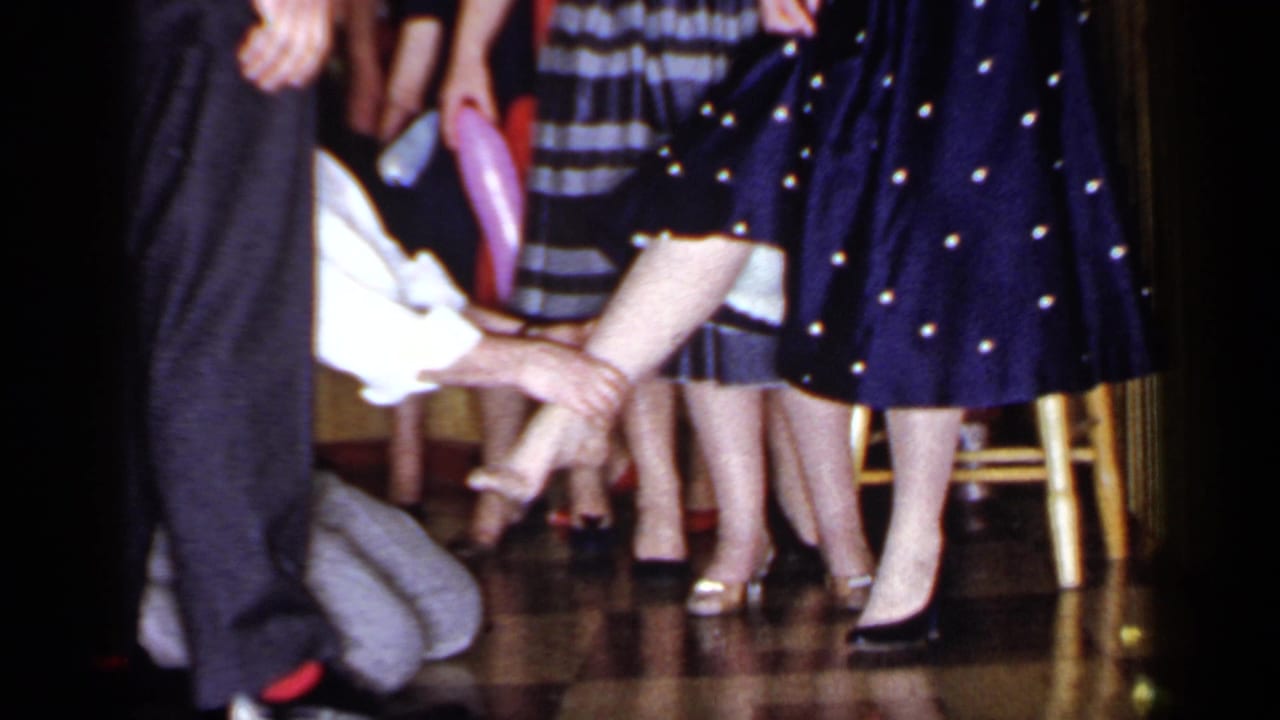 Kneeling man reaches toward a woman's calf while women in dresses stand nearby on a polished floor.