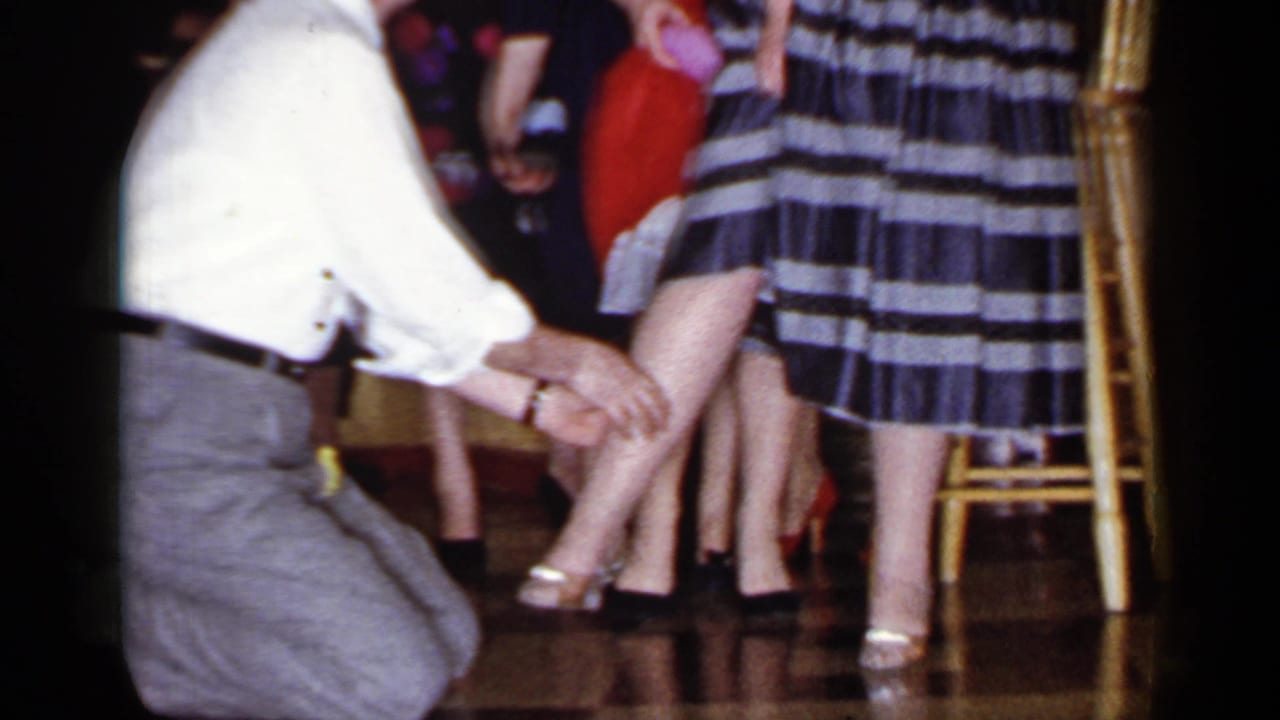 Man kneels beside a line of women in dresses and heels during an indoor gathering.