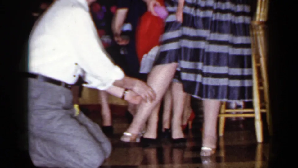 A man in a white shirt and grey trousers kneels beside women in striped and dark dresses at a 1966 Mineola social event.
