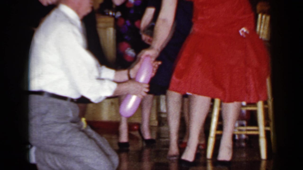 In 1966 Mineola, New York home-movie footage, a blindfolded man kneels near a woman in a red dress as guests stand close by.