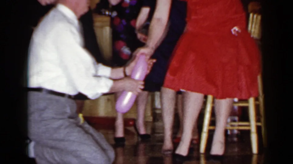 In 1966 Mineola, New York home-movie footage, a blindfolded man kneels near a woman in a red dress as guests stand close by.