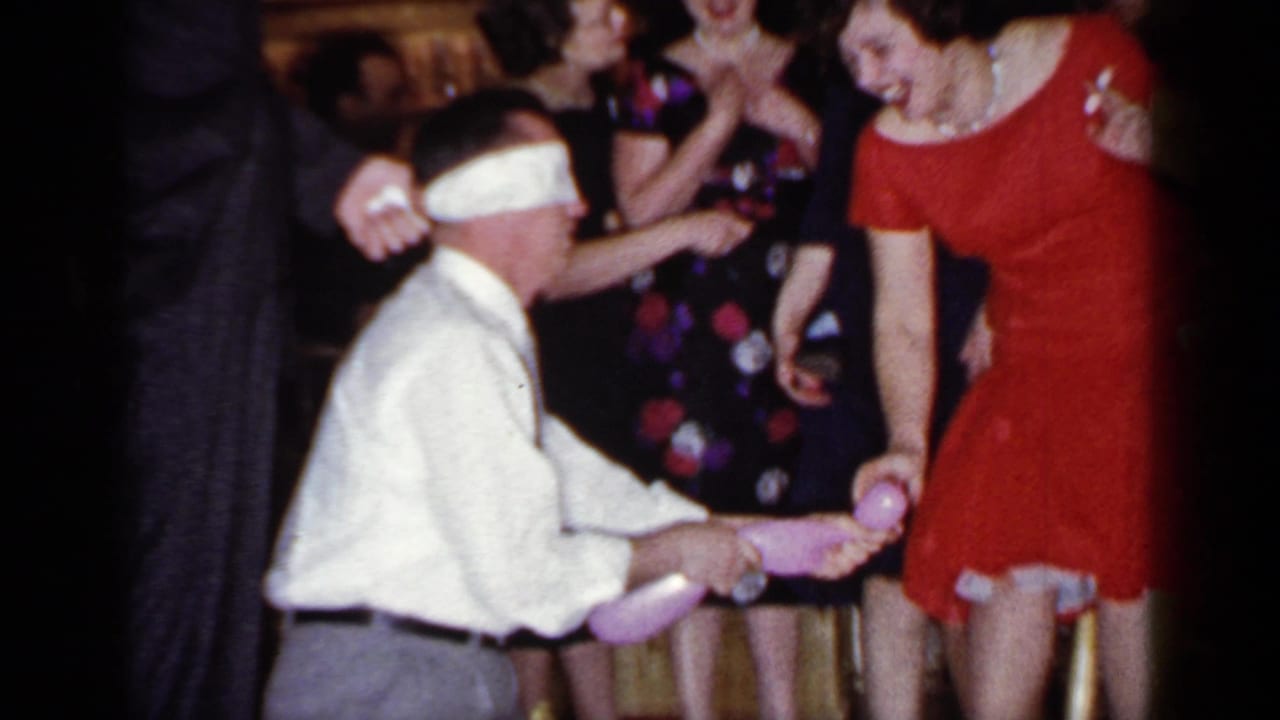Blindfolded man reaches toward a woman in a red dress as guests watch.
