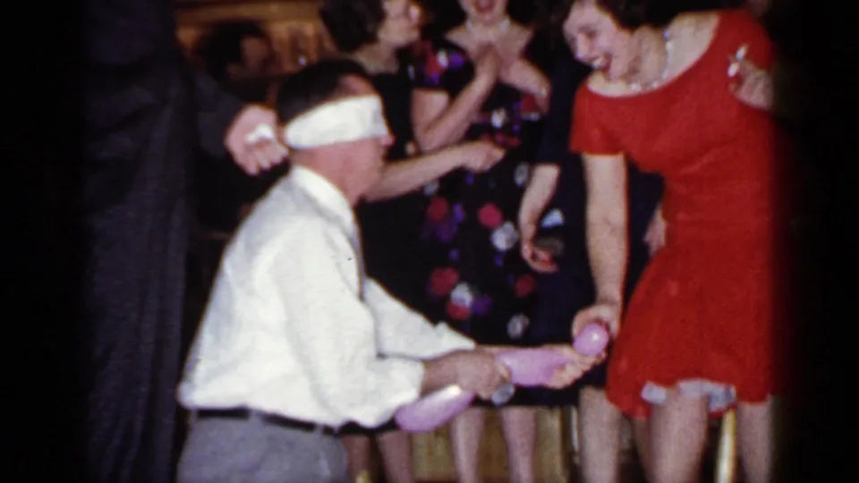The blindfolded man reaches forward at an indoor 1966 Mineola party while a woman in a red dress and nearby guests watch.