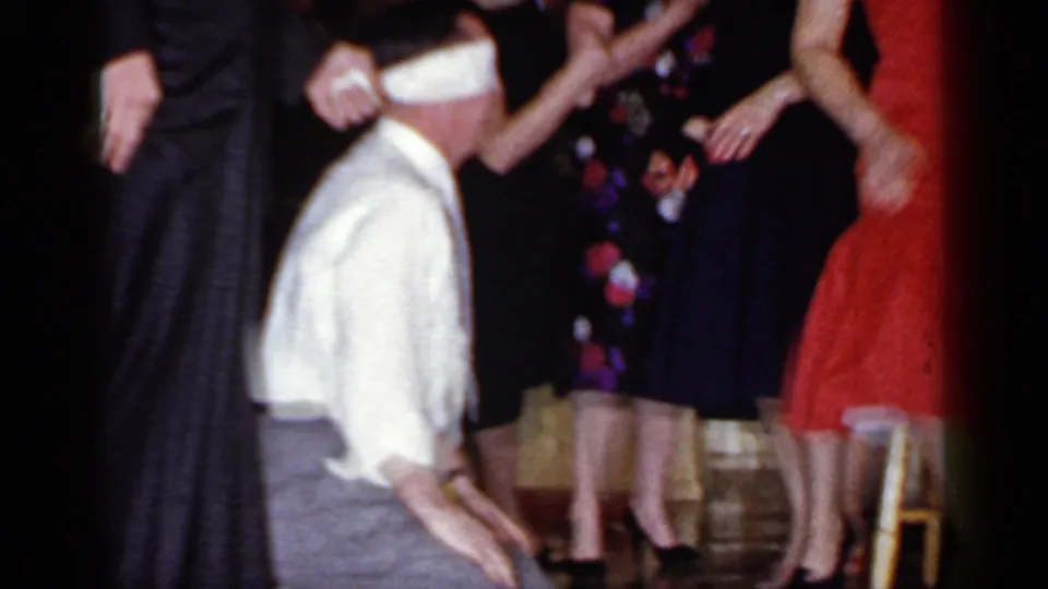 A blindfolded man in a white shirt kneels in a crowded indoor party scene, surrounded by formally dressed guests in 1966 Mineola footage.