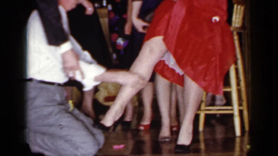 The 1966 Mineola home movie shows the blindfolded man kneeling as a woman in a red dress lifts her leg during the garter game.