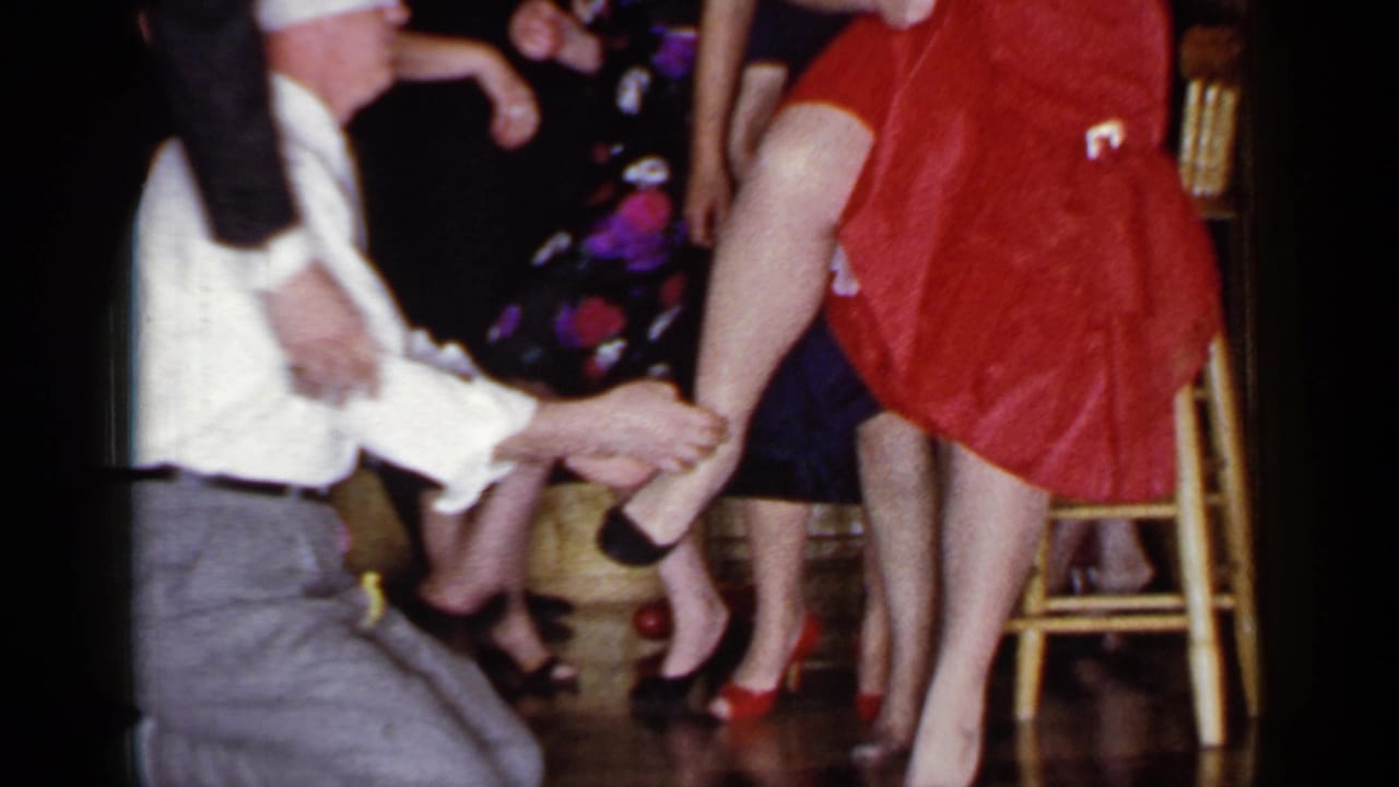 In Mineola, New York, 1966, a kneeling man in a white shirt and cap holds a woman's leg as she stands in a red dress at a party.