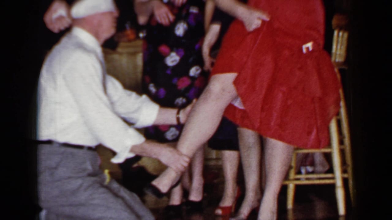 Kneeling man holds a woman's ankle while partygoers stand close behind her.