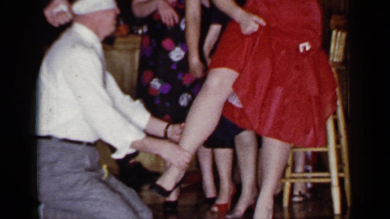 A 1966 Mineola home-movie frame captures the kneeling man holding the red-dressed woman's leg while other adults stand nearby.