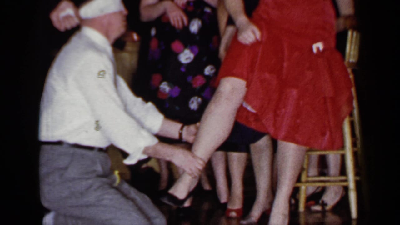 In the Mineola, New York party footage, the kneeling man holds the woman's leg while adults in patterned dresses stand in the background.