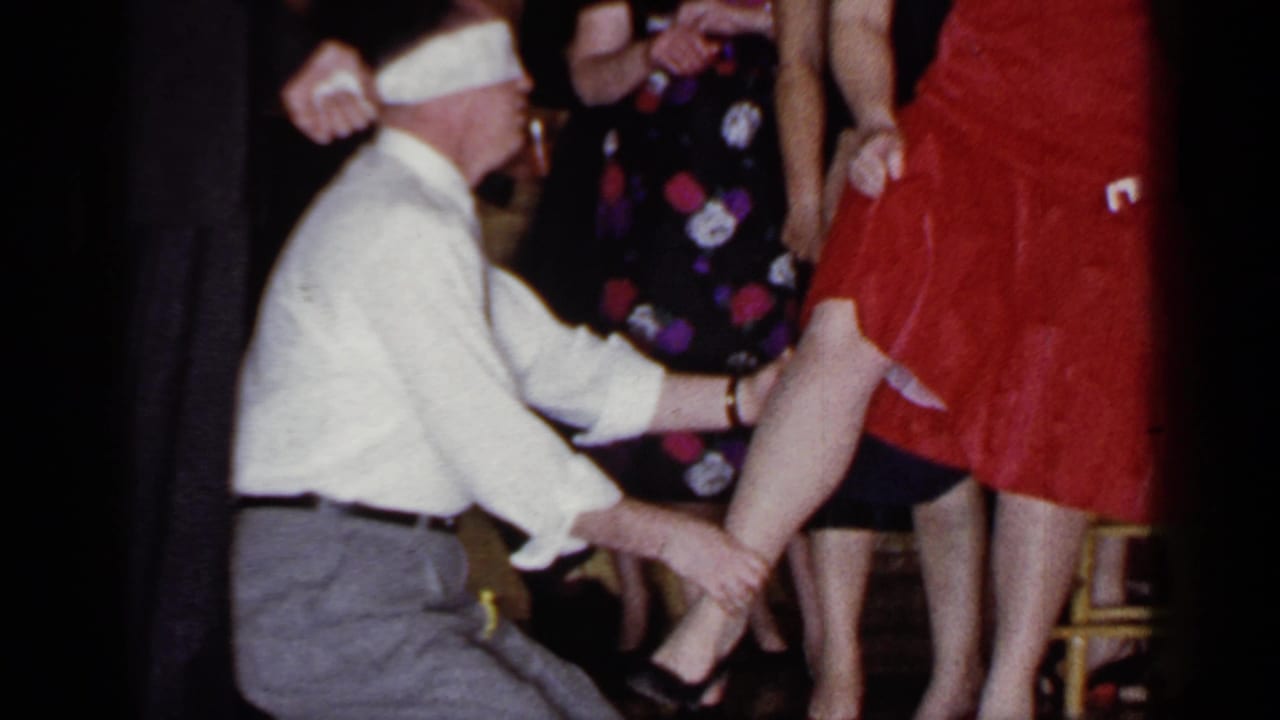 Blindfolded man kneels beside a woman in a red dress while party guests stand nearby.