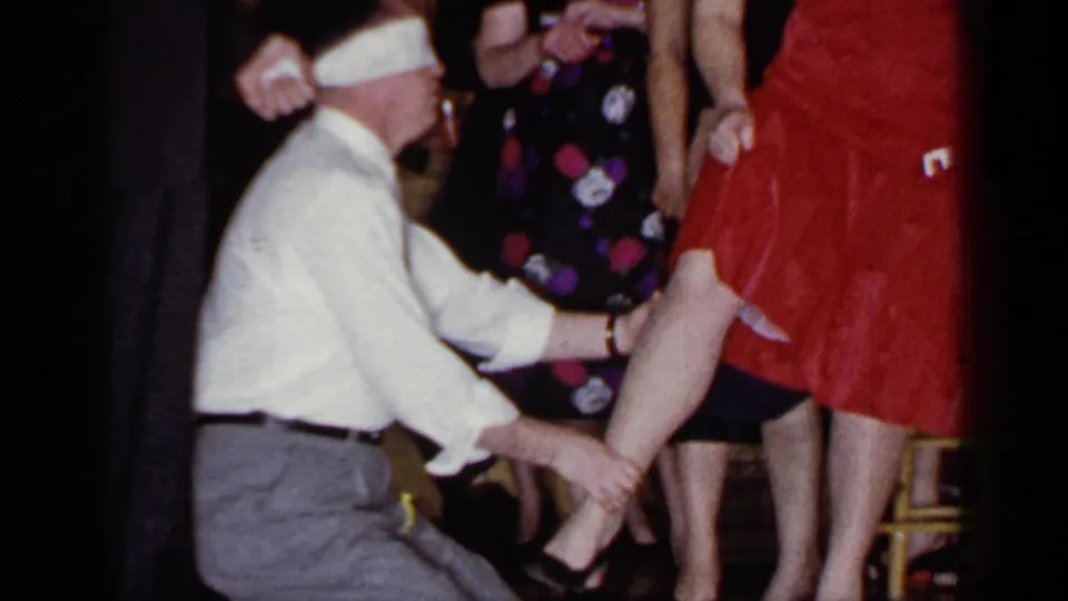 In a 1966 Mineola home-movie party scene, a blindfolded man kneels and touches a woman's leg as guests watch.