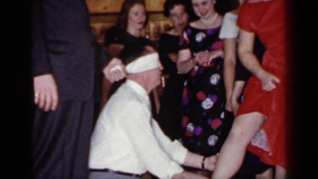 At the Mineola party, the blindfolded kneeling man reaches toward the woman in red while adults in dresses and a suit watch.